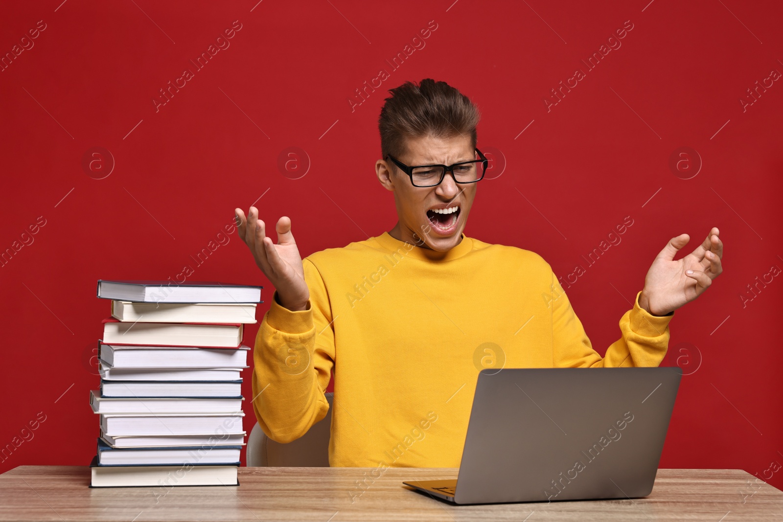 Photo of Emotional student having stress before exam with stack of books and laptop at wooden table against red background