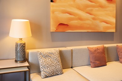 Photo of Comfortable sofa with cushions, beautiful abstract painting and lamp on side table in room