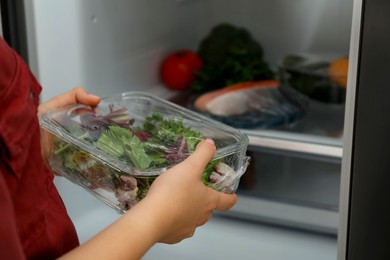 Woman putting glass container of fresh salad with plastic food wrap into refrigerator, closeup