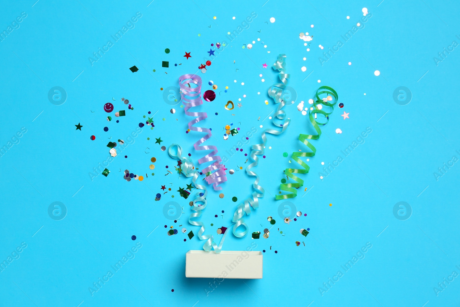 Photo of Shiny confetti and party streamers flying out of little box on light blue background, flat lay