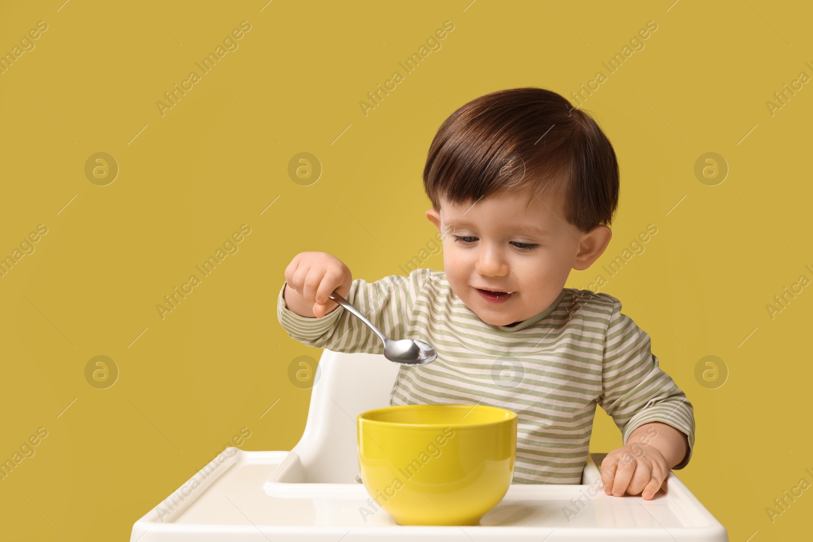 Photo of Cute little kid eating healthy baby food from bowl in high chair on yellow background