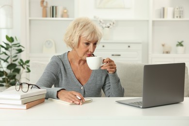 Beautiful senior woman using laptop at white table indoors