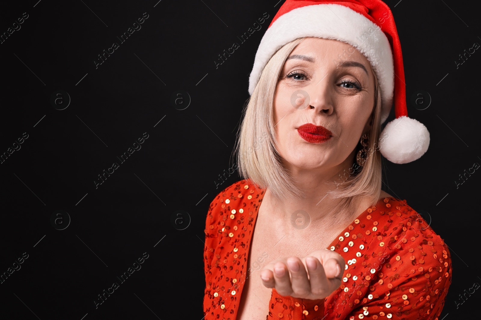 Photo of Woman with perfect makeup in Santa hat sending air kiss on black background, space for text. Christmas celebration