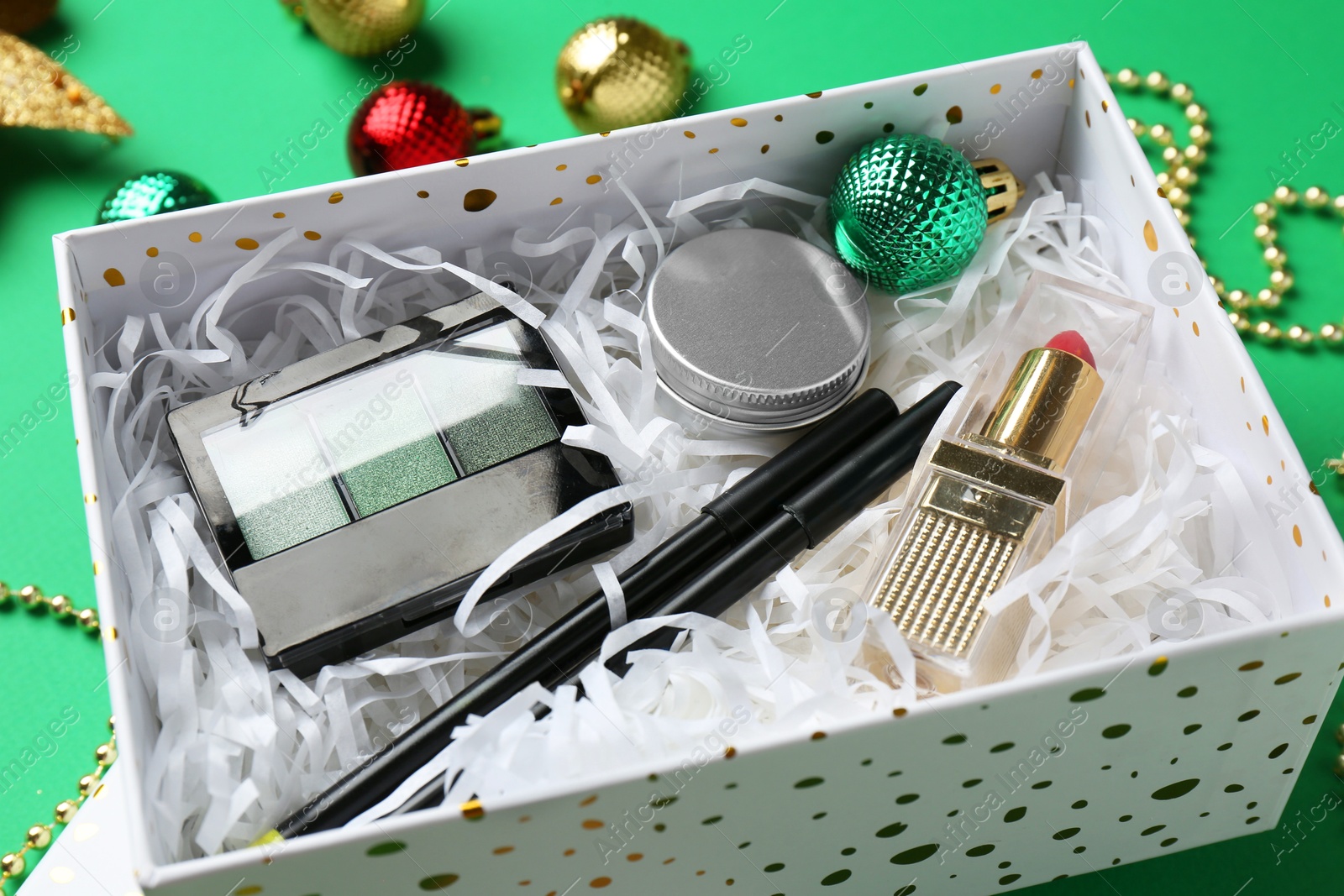 Photo of Christmas gift box with decorative cosmetics and festive ornaments on green background, closeup