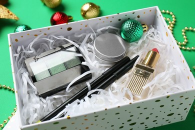 Christmas gift box with decorative cosmetics and festive ornaments on green background, closeup