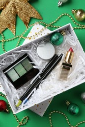 Photo of Christmas gift box with decorative cosmetics and festive ornaments on green background, flat lay