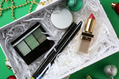 Photo of Christmas gift box with decorative cosmetics and festive ornaments on green background, flat lay