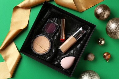 Photo of Christmas present. Decorative cosmetics in gift box and festive balls on green background, flat lay