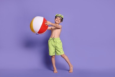 Little boy in beachwear with diving mask and inflatable ball on purple background