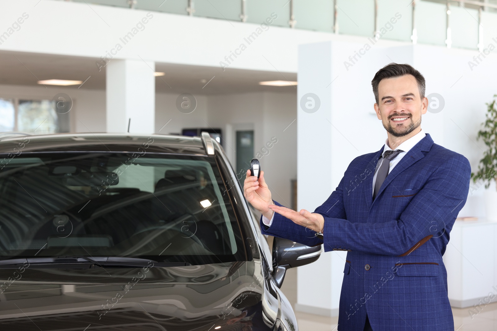 Photo of Happy salesman holding key near new black car in salon