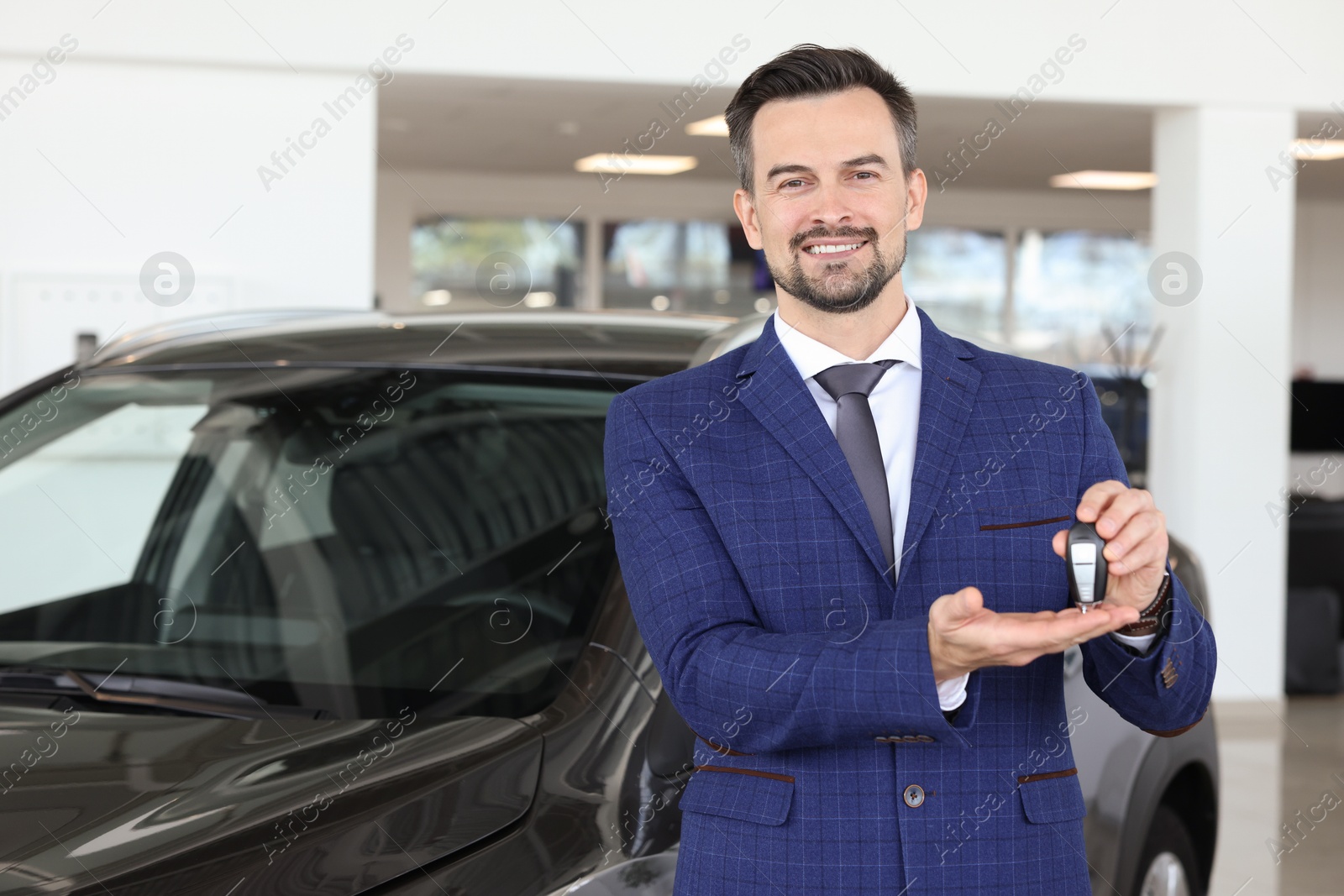 Photo of Happy salesman holding key near new black car in salon