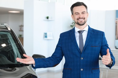 Photo of Happy salesman showing thumbs up near new black car in salon