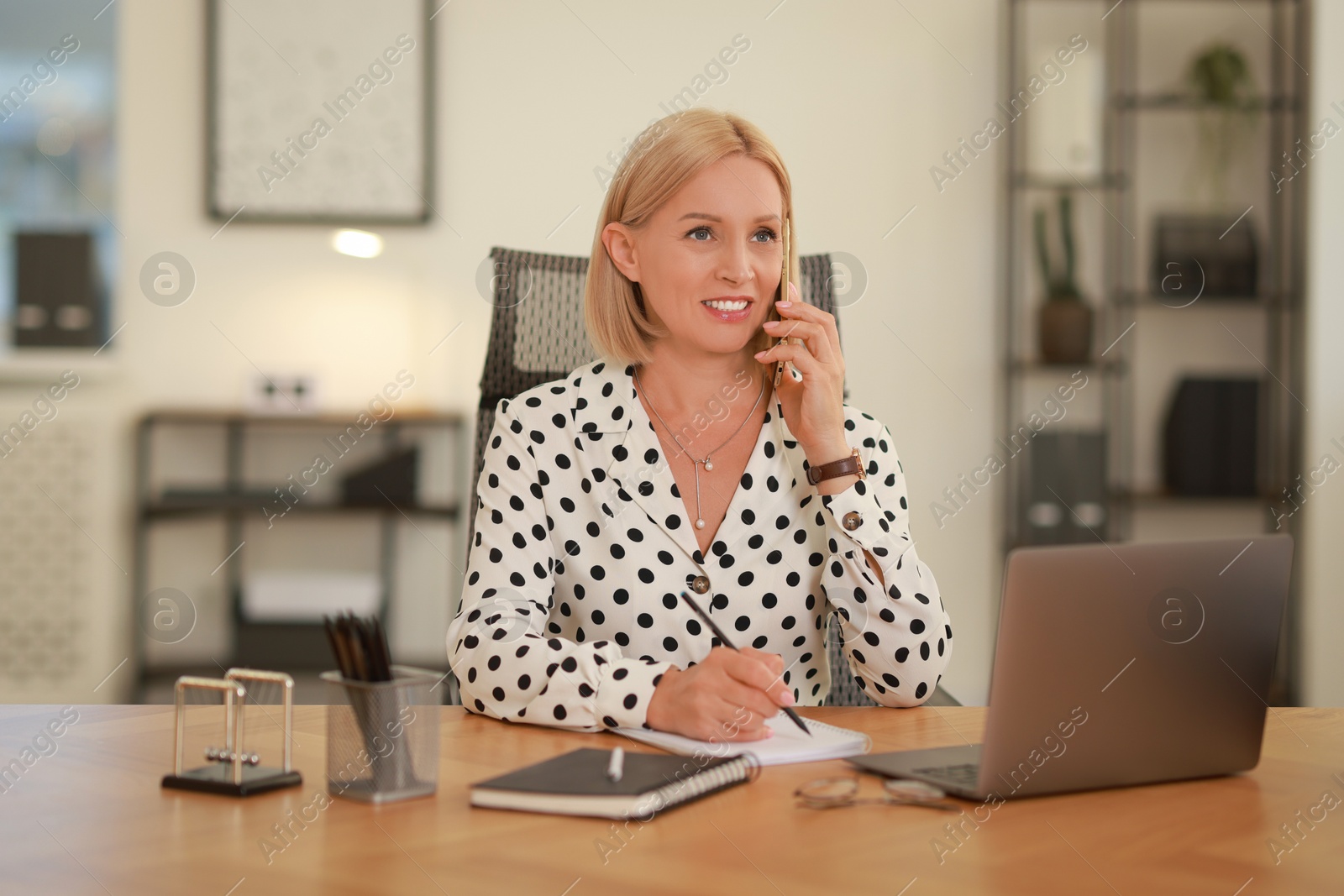 Photo of Smiling middle aged woman talking by smartphone at table in office