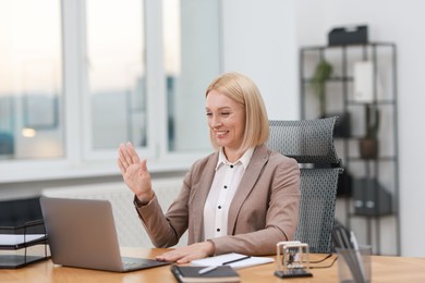 Photo of Smiling middle aged woman having videochat by laptop at table in office