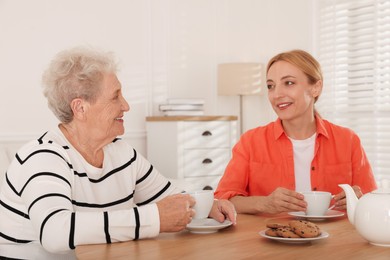 Photo of Caregiver and senior woman enjoying hot drink at table indoors