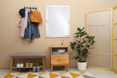 Coat rack with clothes, mirror, houseplant, shoe storage bench and folding screen in hallway. Interior design