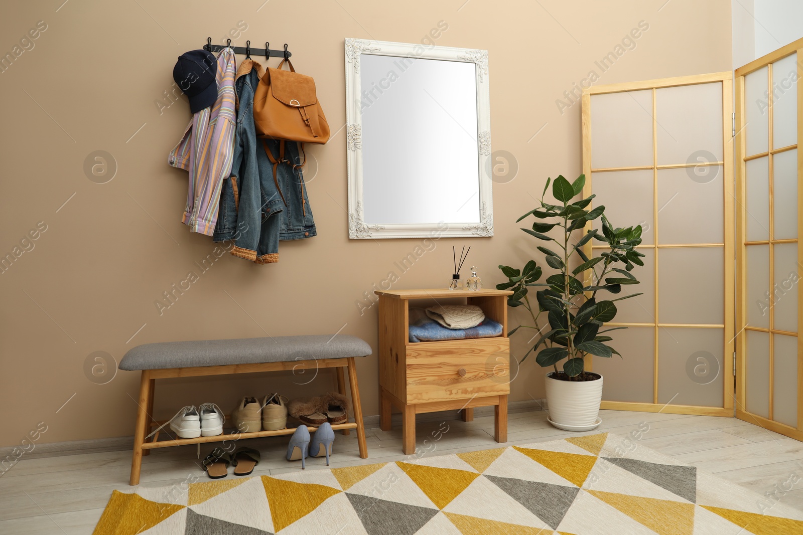 Photo of Coat rack with clothes, mirror, houseplant, shoe storage bench and folding screen in hallway. Interior design