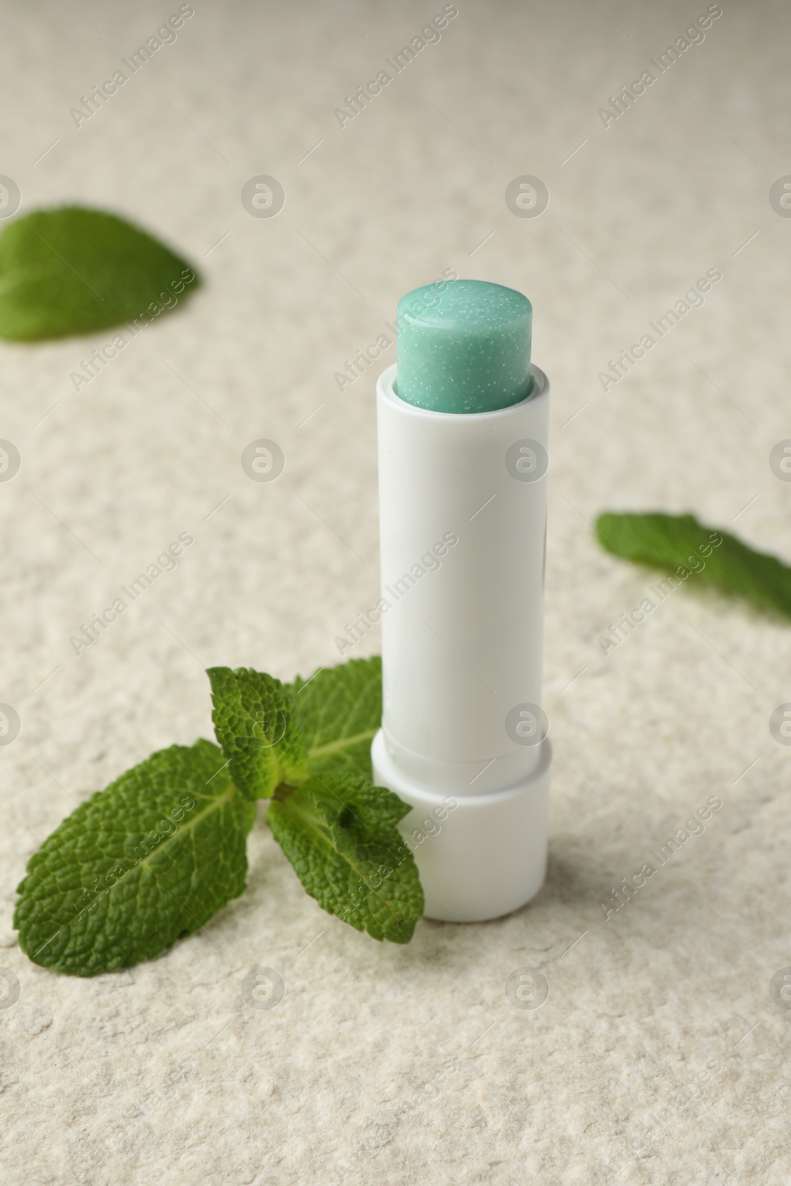 Photo of Mint lip balm and green leaves on beige textured table, closeup. Cosmetic product