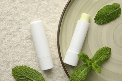 Photo of Mint lip balms and green leaves on beige textured table, top view. Cosmetic product