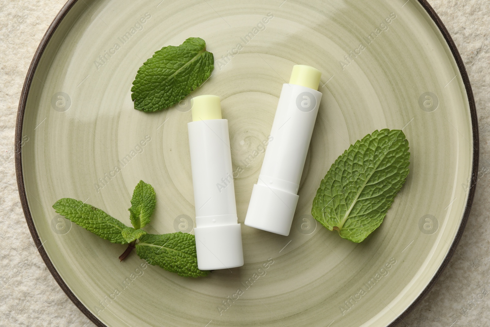 Photo of Mint lip balms and green leaves on beige textured table, top view. Cosmetic product