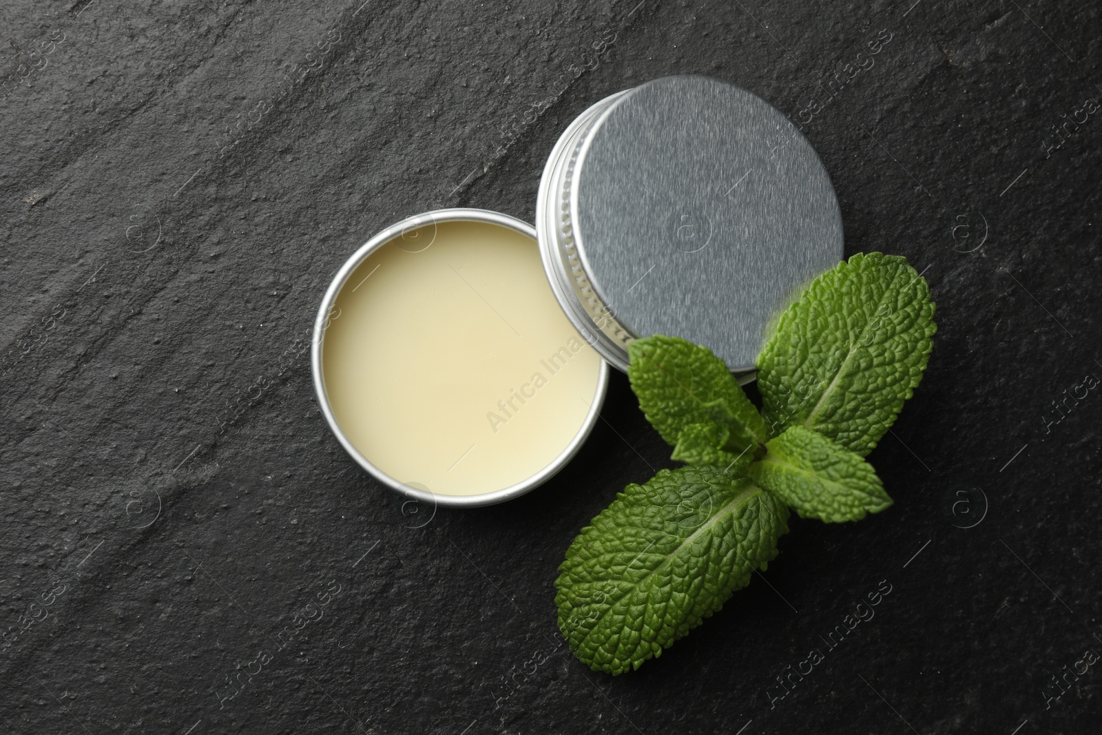 Photo of Mint lip balm and green leaves on black table, top view. Cosmetic product