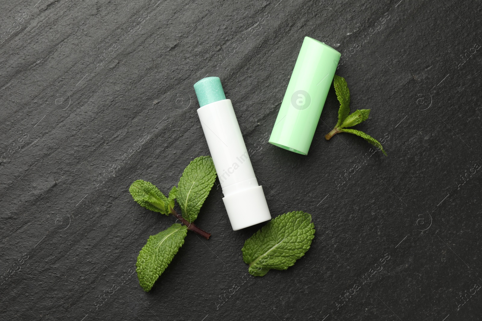 Photo of Mint lip balm and green leaves on black table, flat lay. Cosmetic product