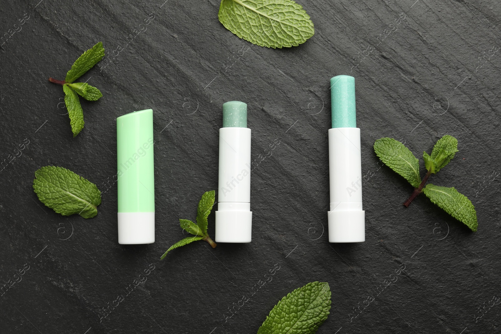 Photo of Mint lip balms and green leaves on black table, flat lay. Cosmetic product