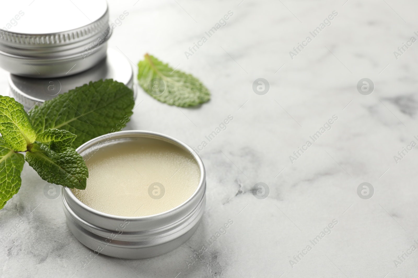 Photo of Mint lip balms and green leaves on white marble table, closeup. Space for text