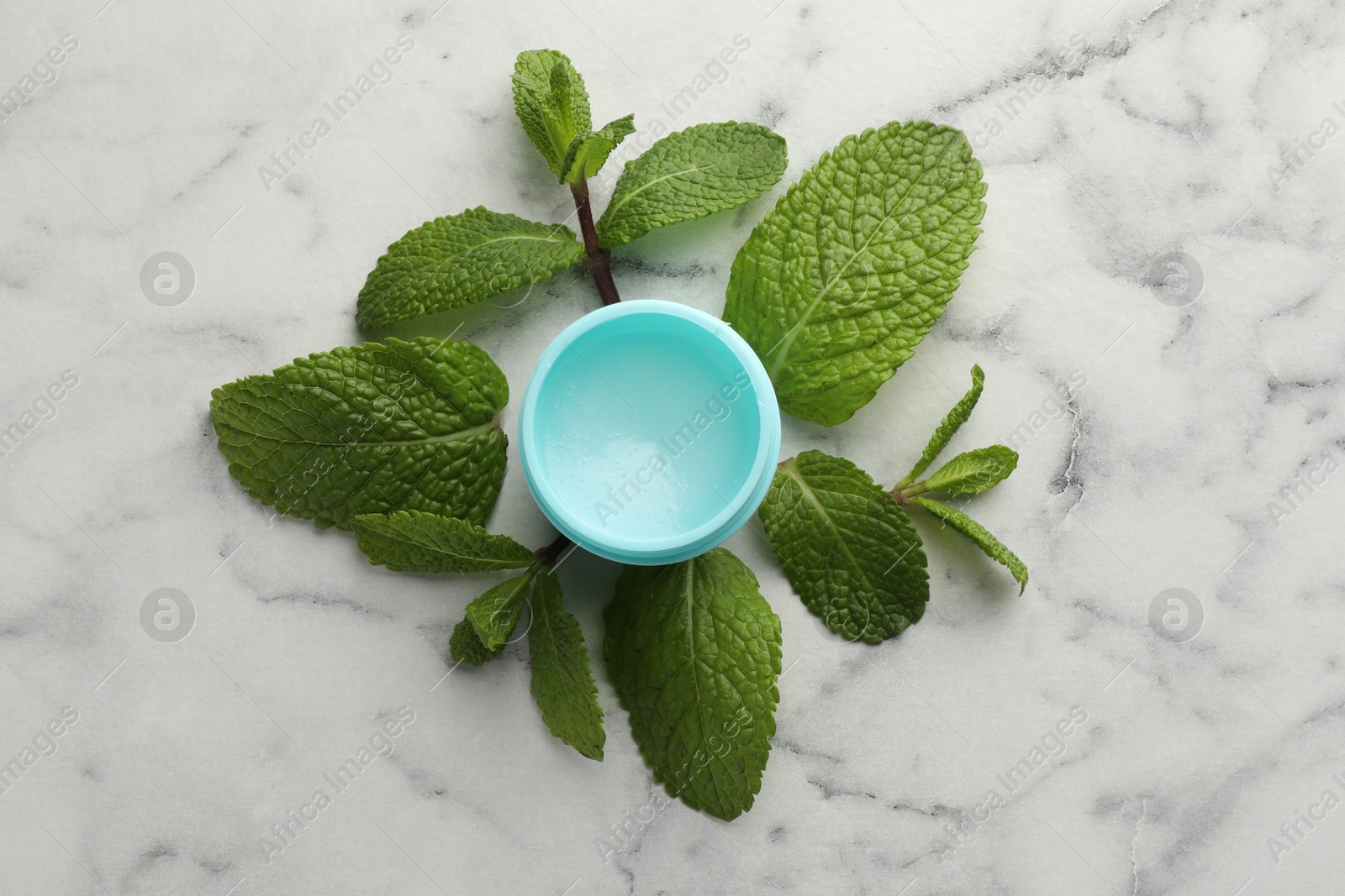 Photo of Mint lip balm and green leaves on white marble table, top view. Cosmetic product