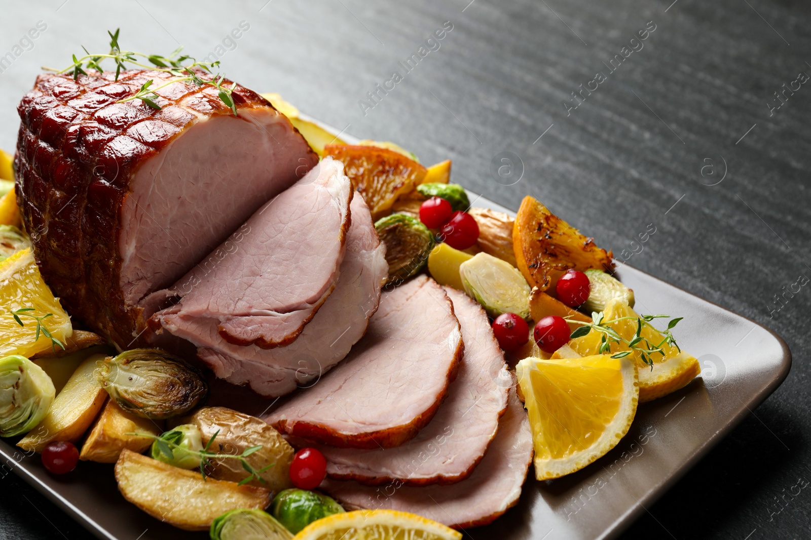 Photo of Tasty baked ham served with vegetables and oranges on black table, closeup