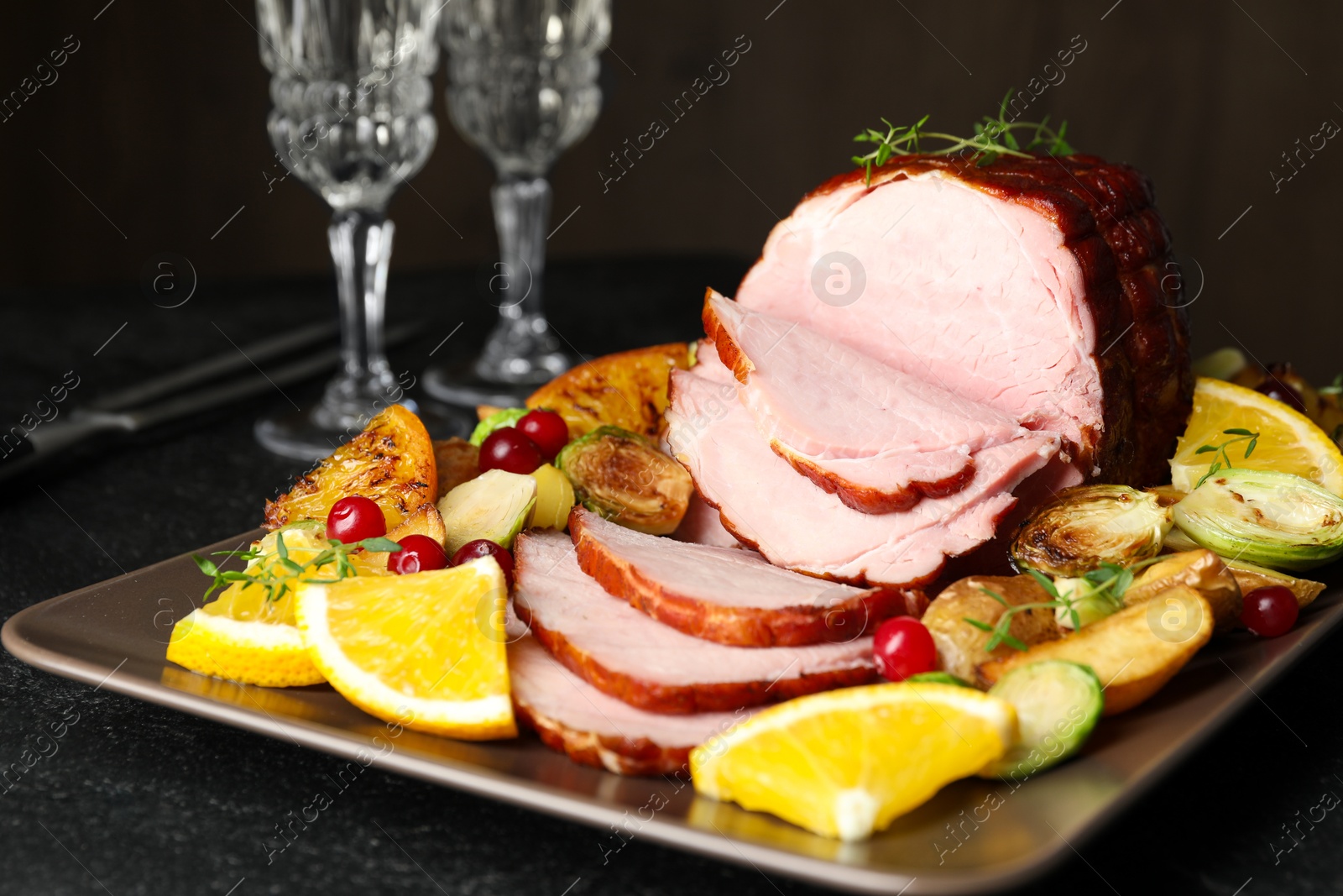 Photo of Tasty baked ham served with vegetables and oranges on black table, closeup