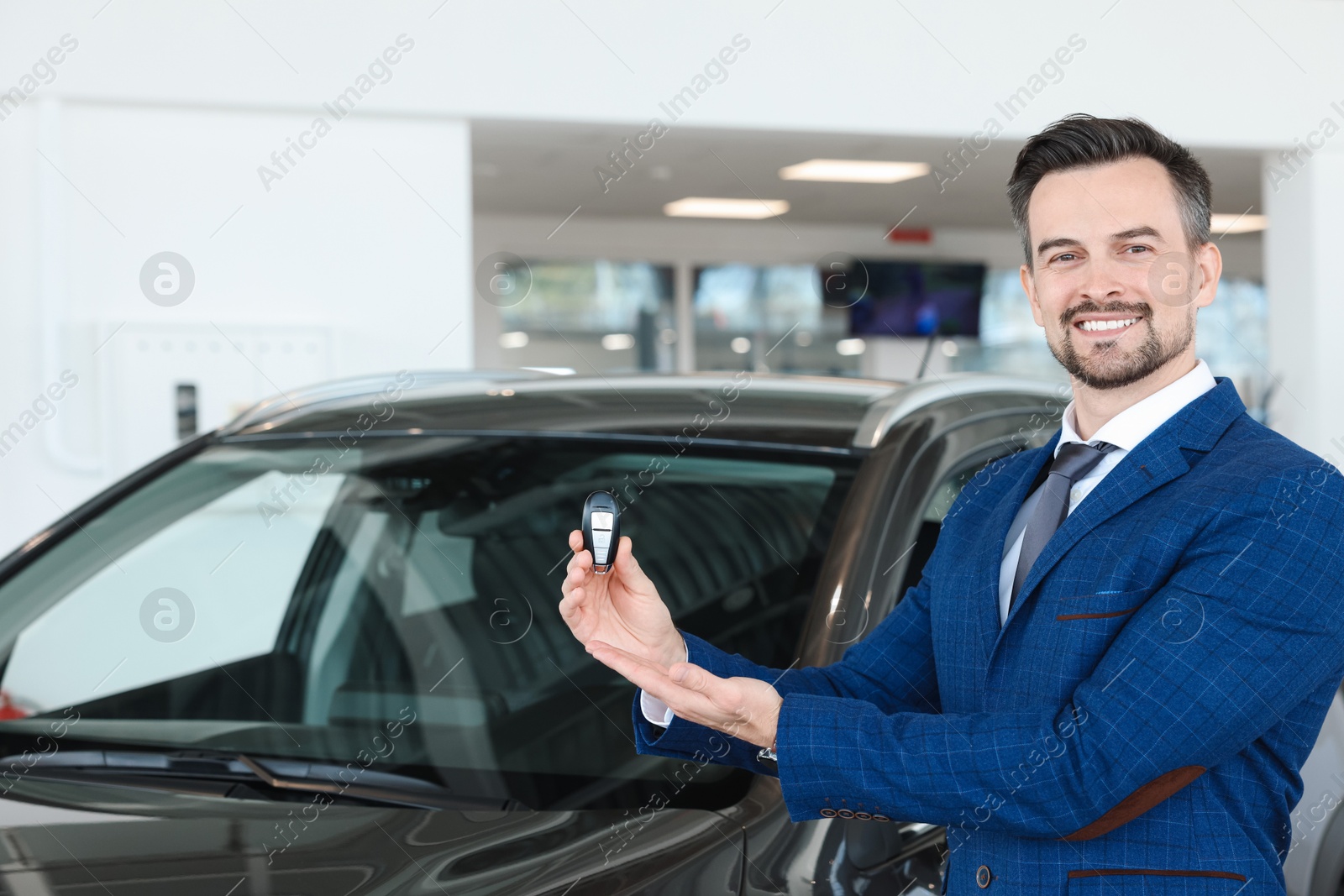 Photo of Happy salesman holding key near new black car in salon