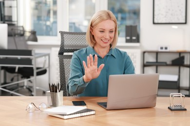 Photo of Smiling middle aged woman having videochat by laptop at table in office