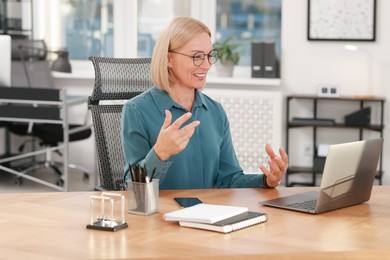 Smiling middle aged woman having videochat by laptop at table in office