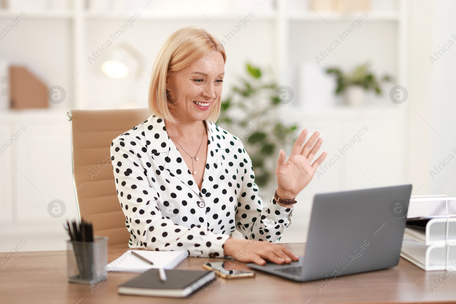 Photo of Smiling middle aged woman having videochat by laptop at table in office