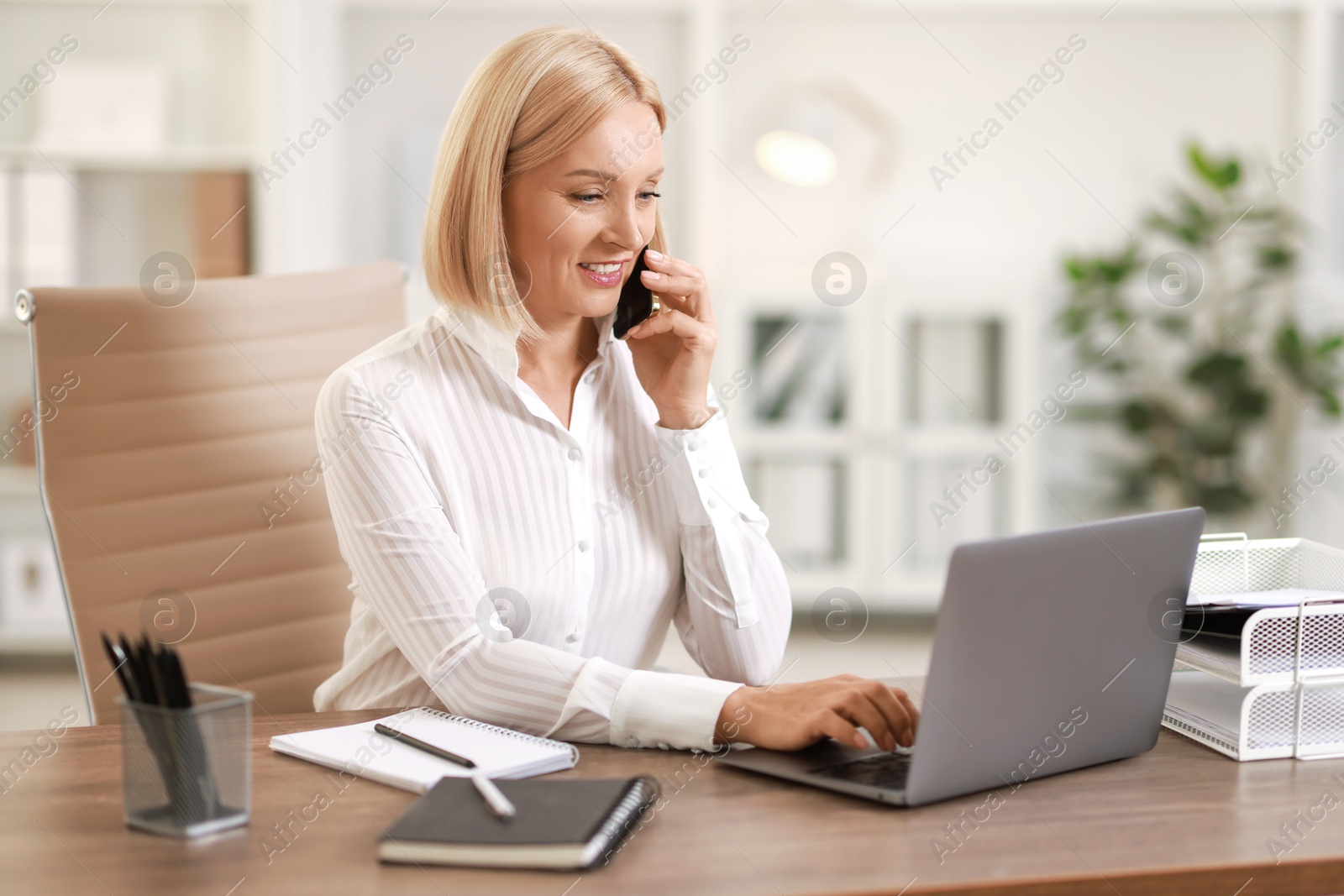 Photo of Smiling middle aged woman talking by smartphone at table in office