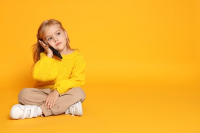 Photo of Cute little girl talking on smartphone against orange background, space for text