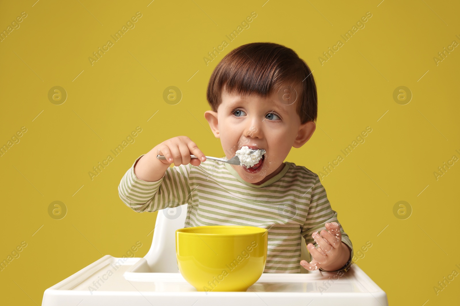 Photo of Cute little kid eating healthy baby food from bowl in high chair on yellow background