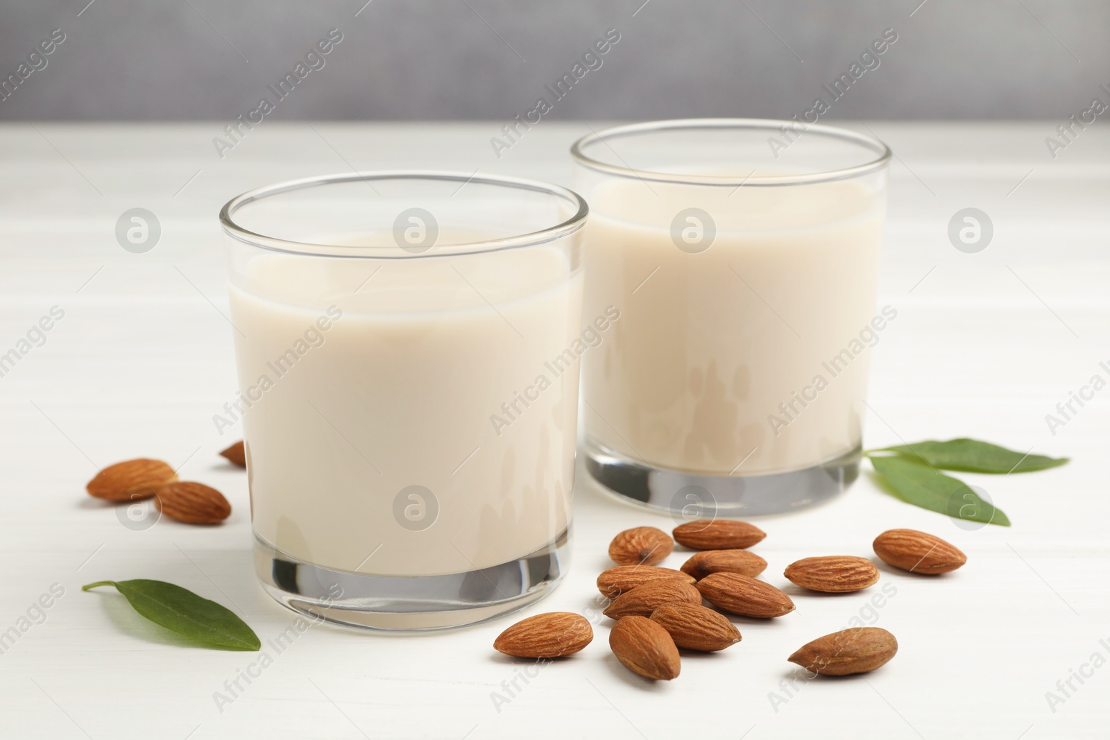 Photo of Fresh nut milk in glasses, green leaves and almonds on white wooden table, closeup