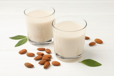 Fresh nut milk in glasses, green leaves and almonds on white wooden table, closeup