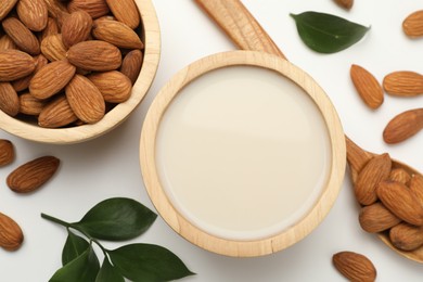 Fresh nut milk in bowl, almonds and green leaves on white table, flat lay