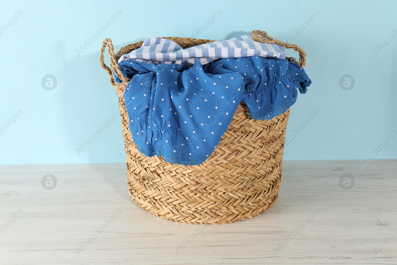 Photo of Wicker basket full of laundry on wooden table against light blue background