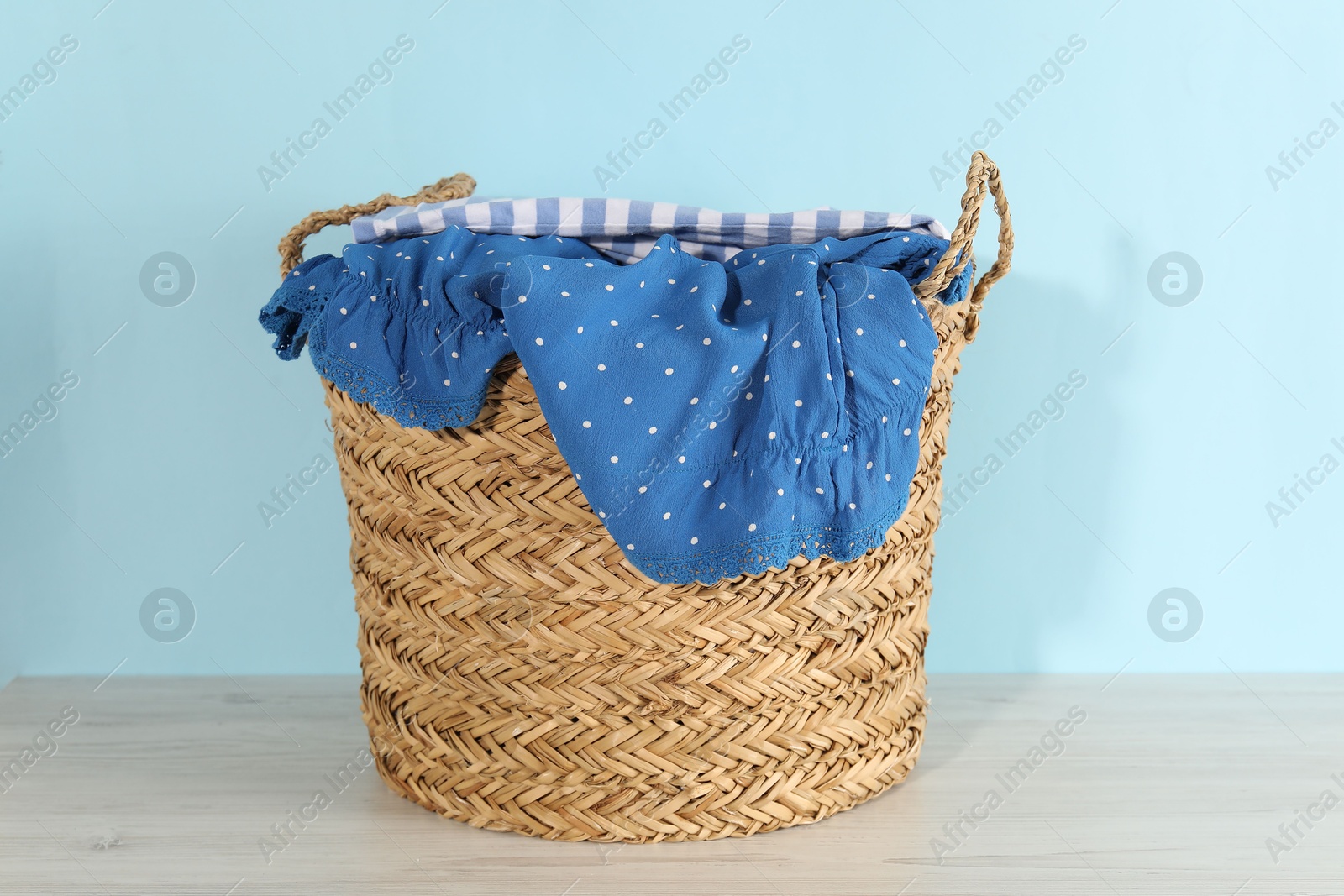 Photo of Wicker basket full of laundry on wooden table against light blue background