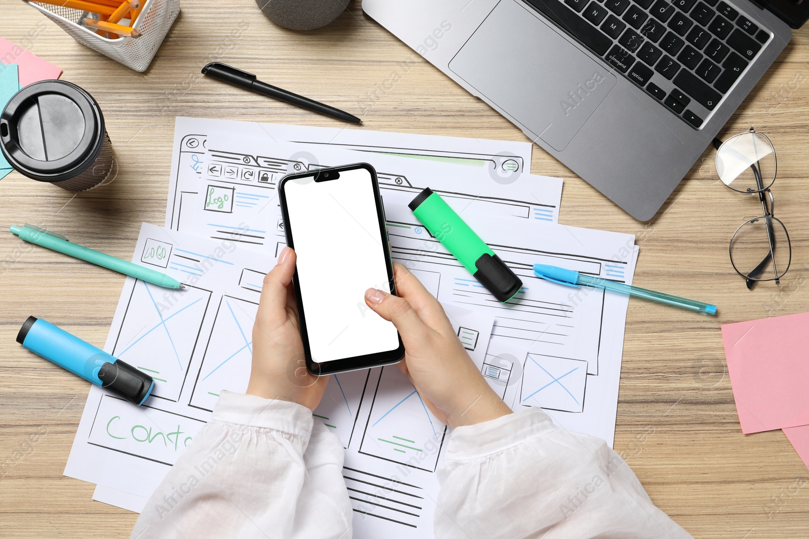 Photo of Website designer using smartphone at wooden table with wireframes and laptop, top view