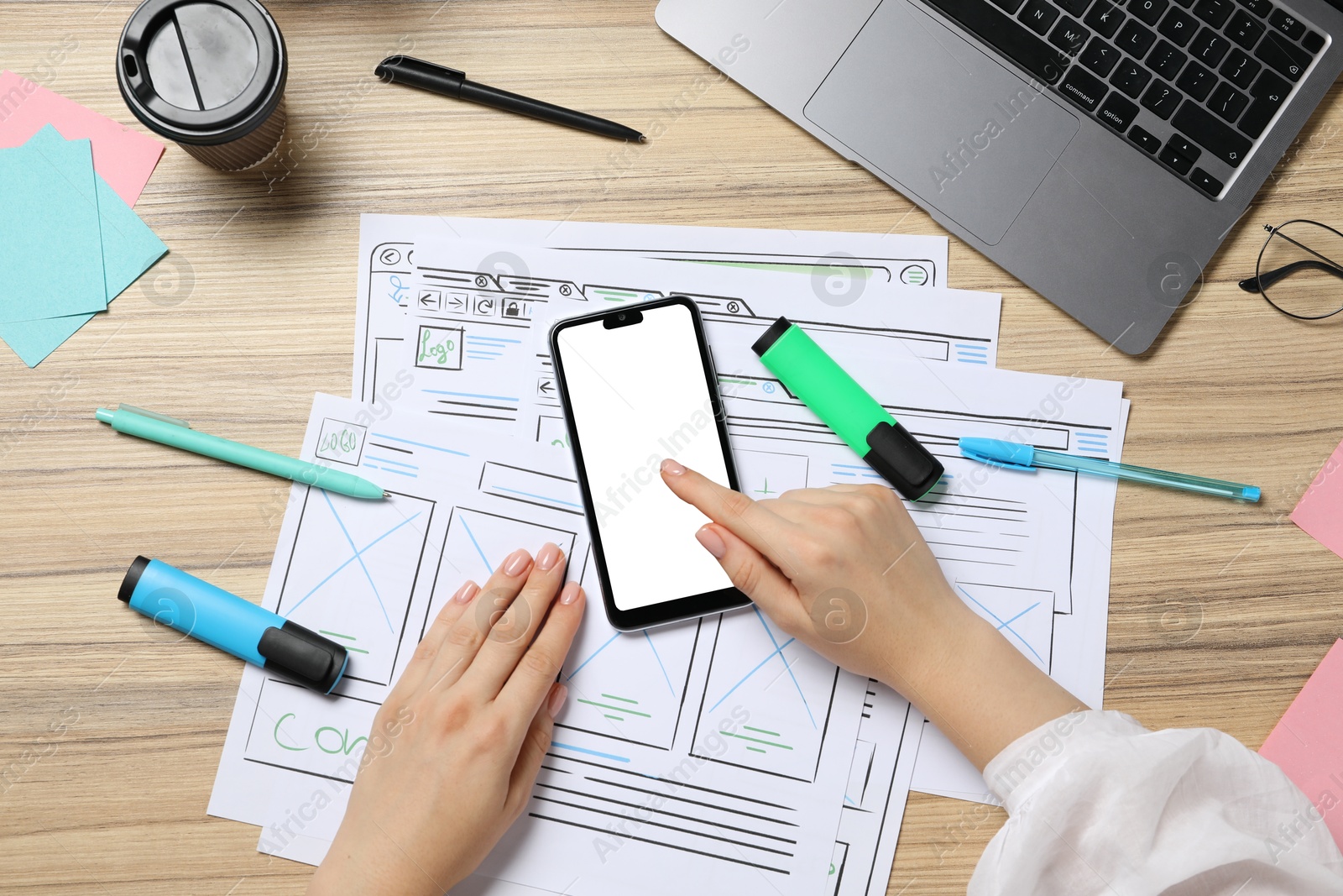 Photo of Website designer using smartphone at wooden table with wireframes and laptop, top view