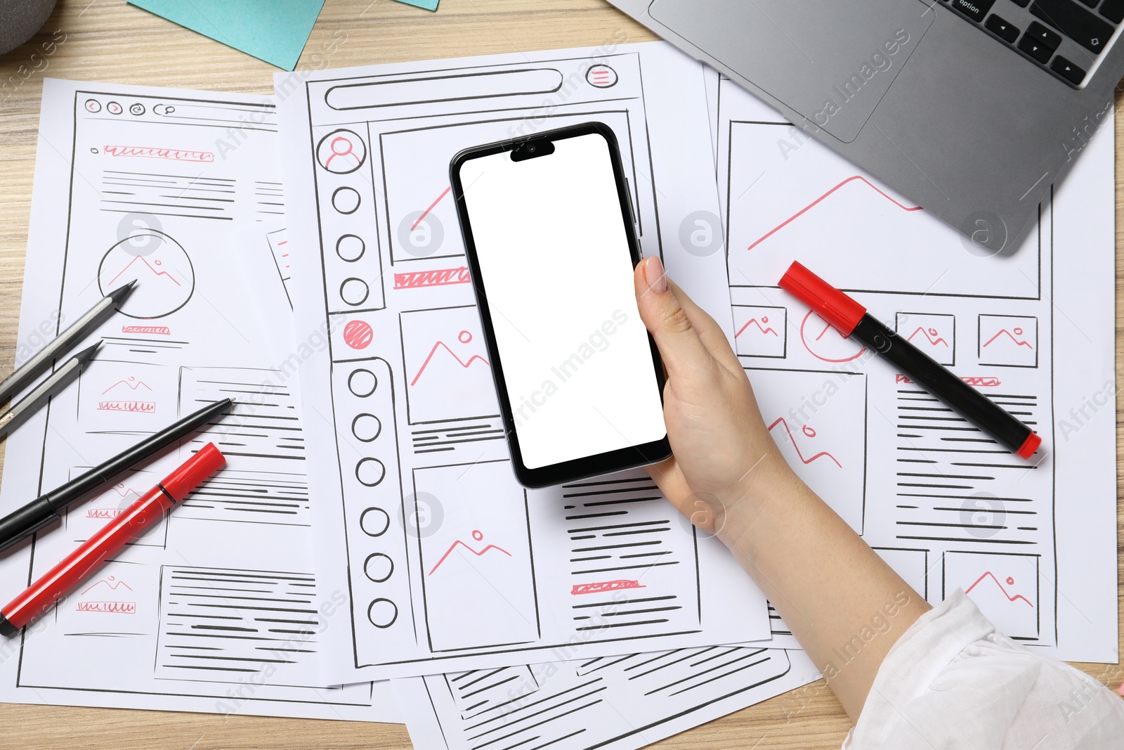 Photo of Website designer holding smartphone at wooden table with wireframes and stationery, top view