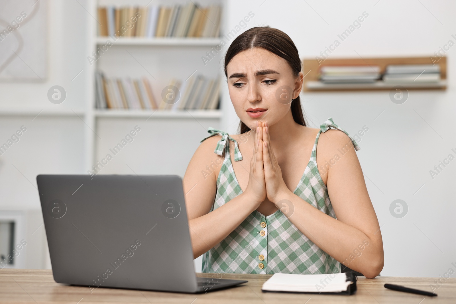 Photo of Worried student praying for good exam result at table with laptop indoors