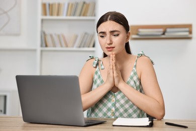 Worried student praying for good exam result at table with laptop indoors