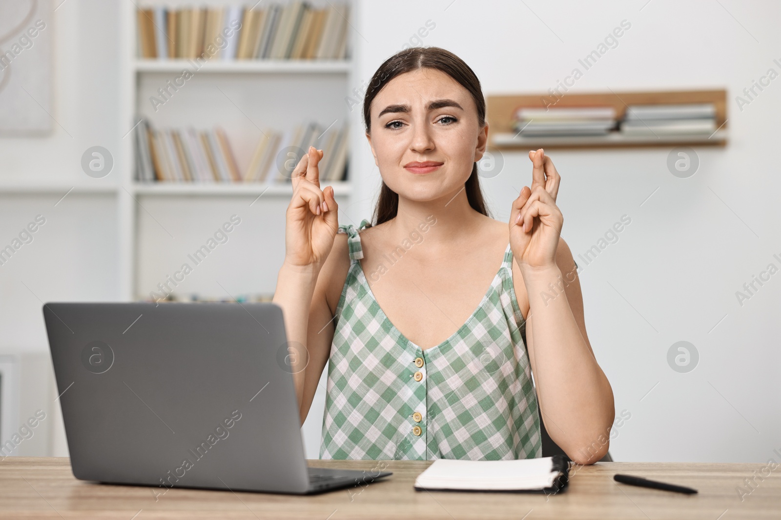 Photo of Worried student with crossed fingers at table with laptop indoors. Hope for good exam result