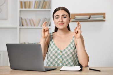 Worried student with crossed fingers at table with laptop indoors. Hope for good exam result